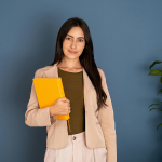 Confident young woman standing against a blue wall, holding a yellow folder — representing creative mums starting a home-based business.”